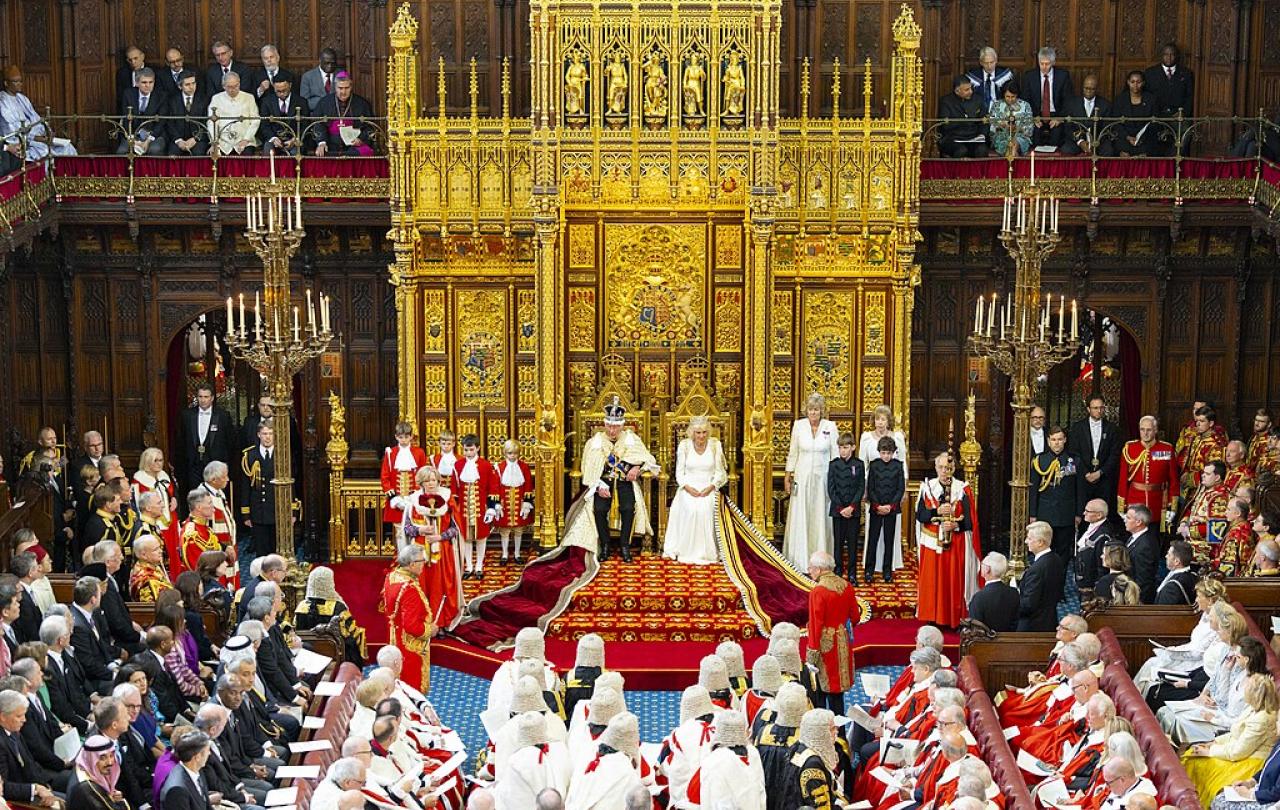 A wide angle picture shows a king and queen on thrones before many people in ceremonial clothes.