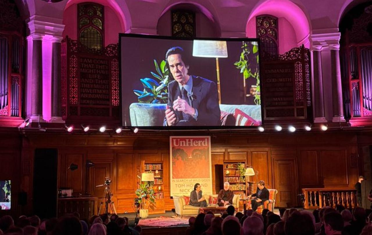 A view of an in-conversation on a stage, with a video screen above showing a close up.