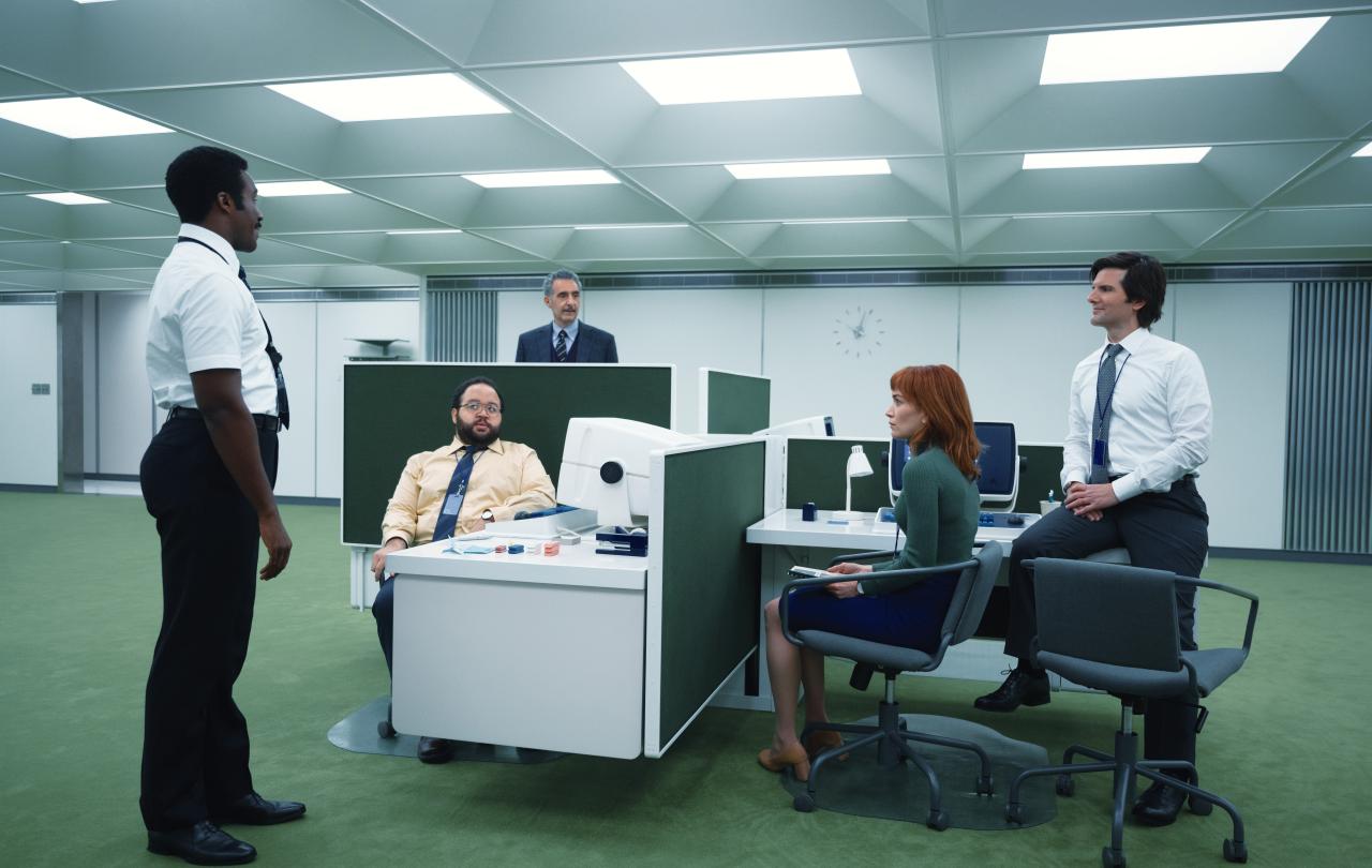 In a retro-future styled office, workers stand or sit around a pod of desks.