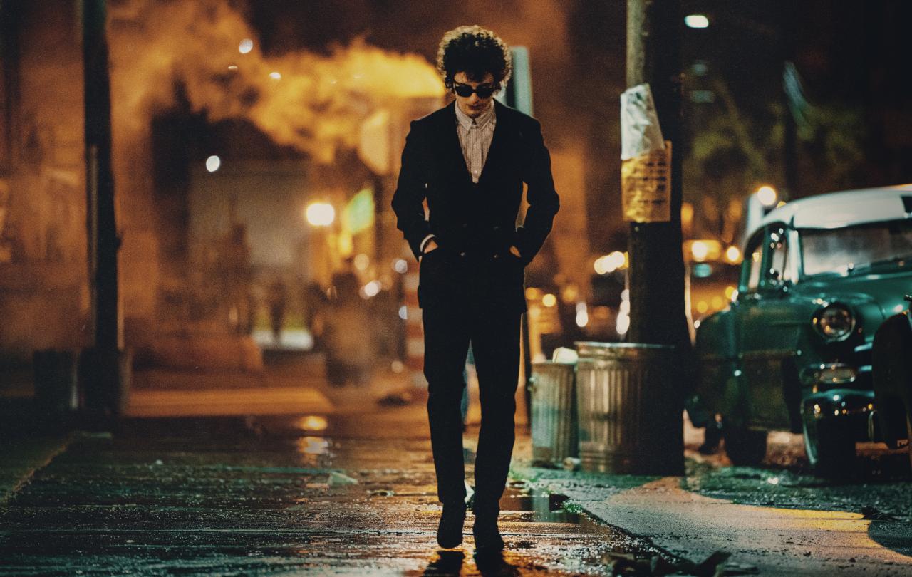 A hunched man wearing dark glasses and a dark suit, walks down a New York Street at night, with his hands in his pockets.