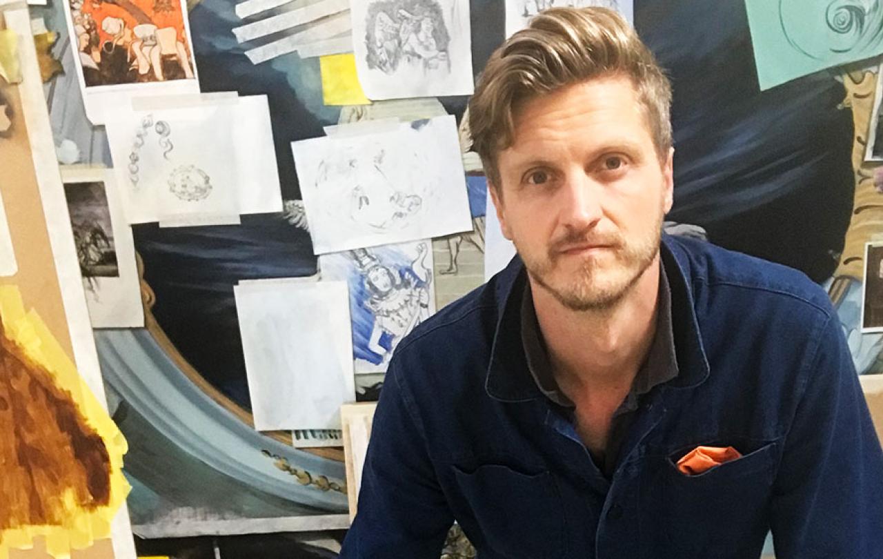 An artist sits in front of a board covered in images, canvases and paper.