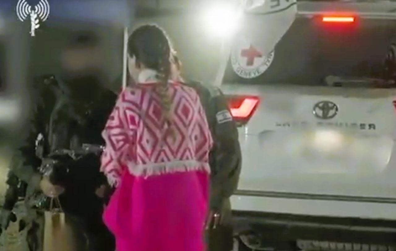 At a night time hostage exchange, a hostage wears a pink top and is guide toward the side of a white vehicle bearing a small red cross.