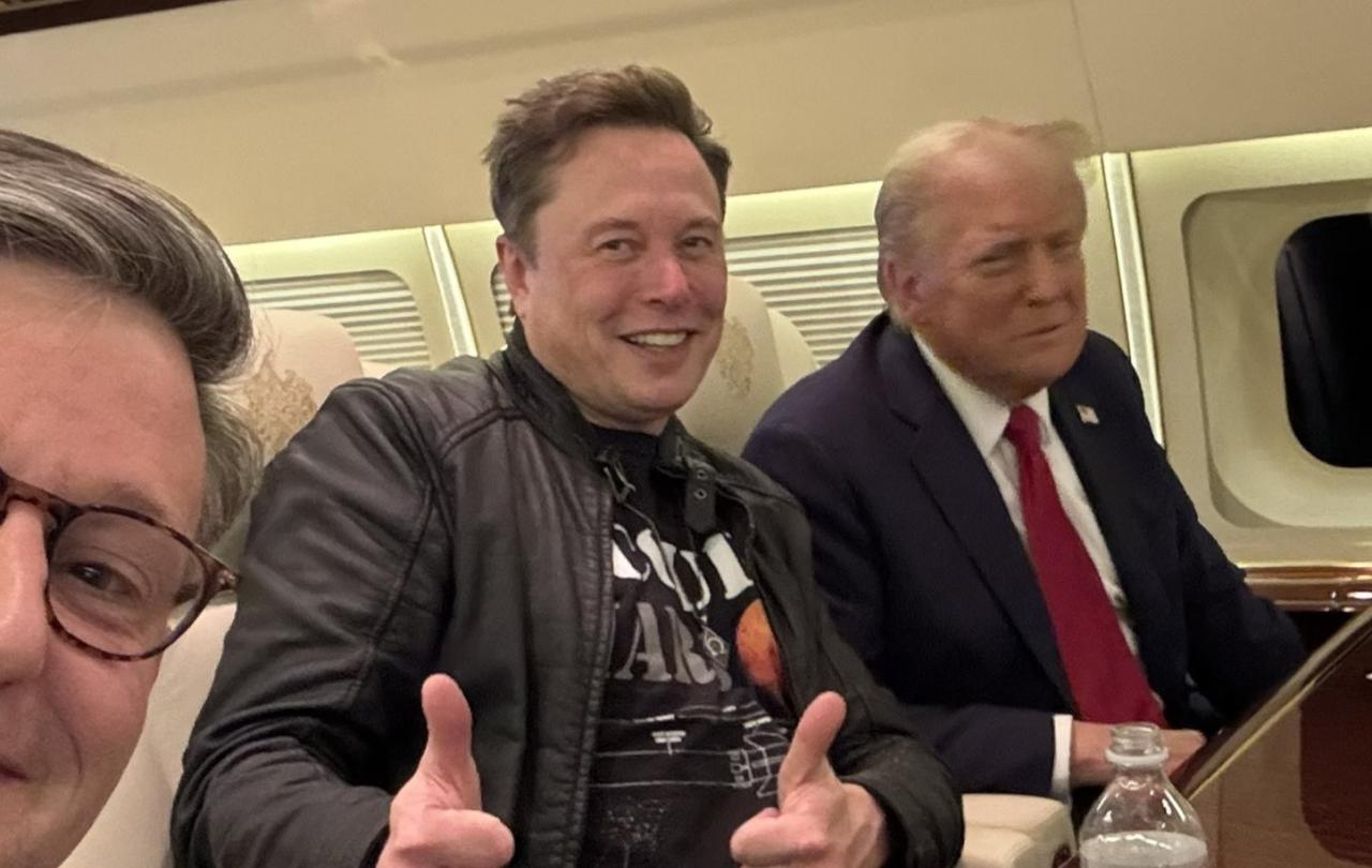 Elon Musk sits next to Donald Trump on a plan, while giving the thumbs up gesture with both hands.