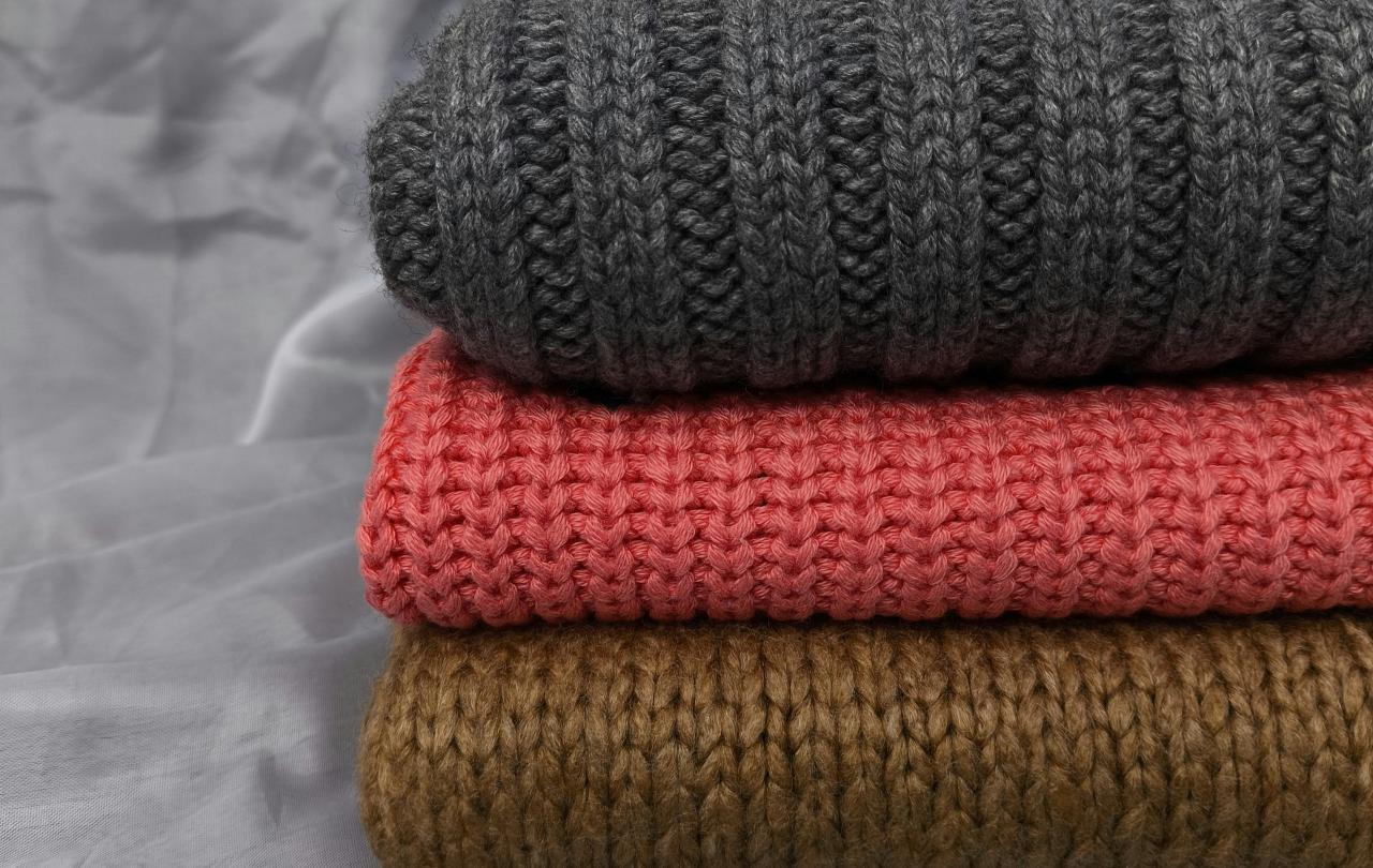 A pile of folded knitted jumpers sit on a grey back ground, one is also grey, the others are red and brown.