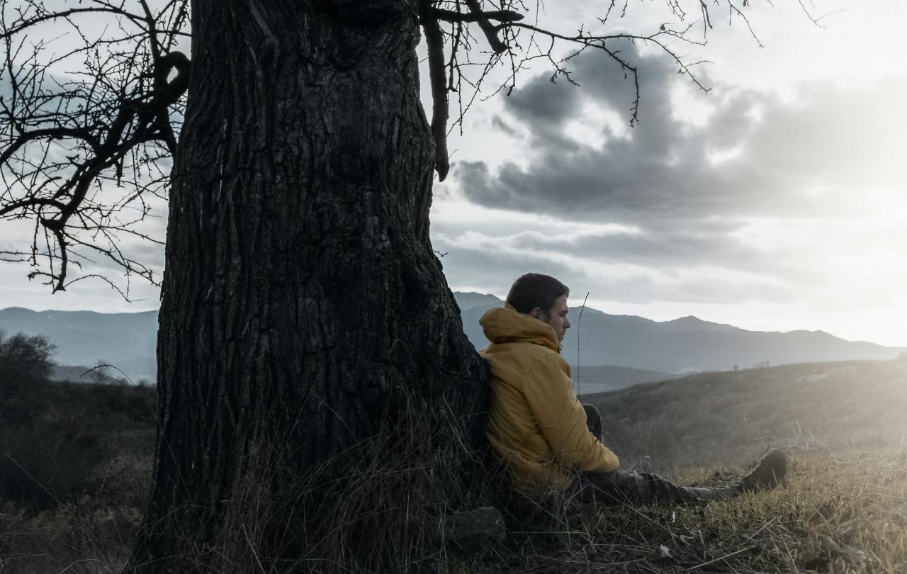 A wrapped-up man sits and leans against a bare tree, as dark clouds give way to sun.