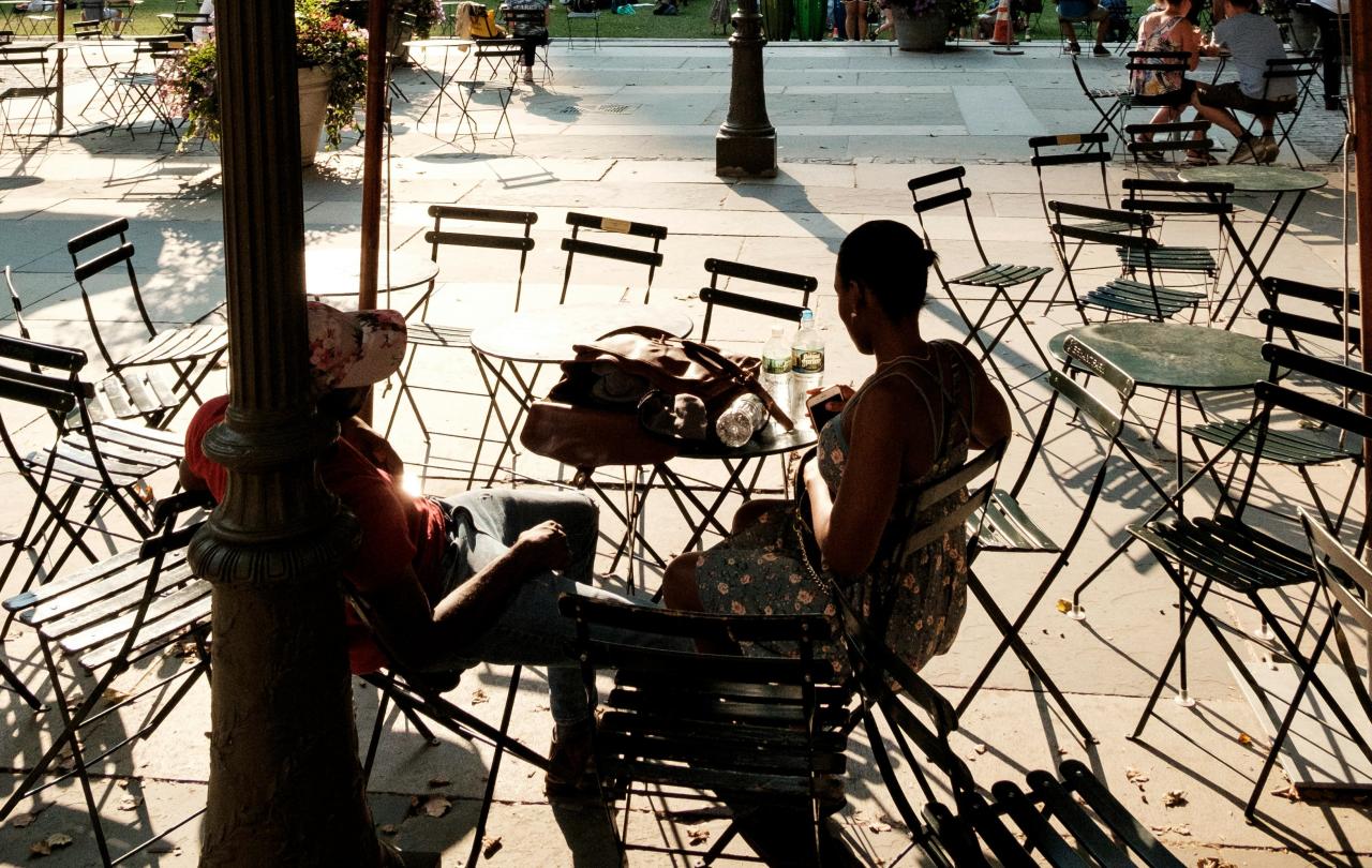 Two people. sitting at a street cafe amid empty tables and chairs, are silhoutted.