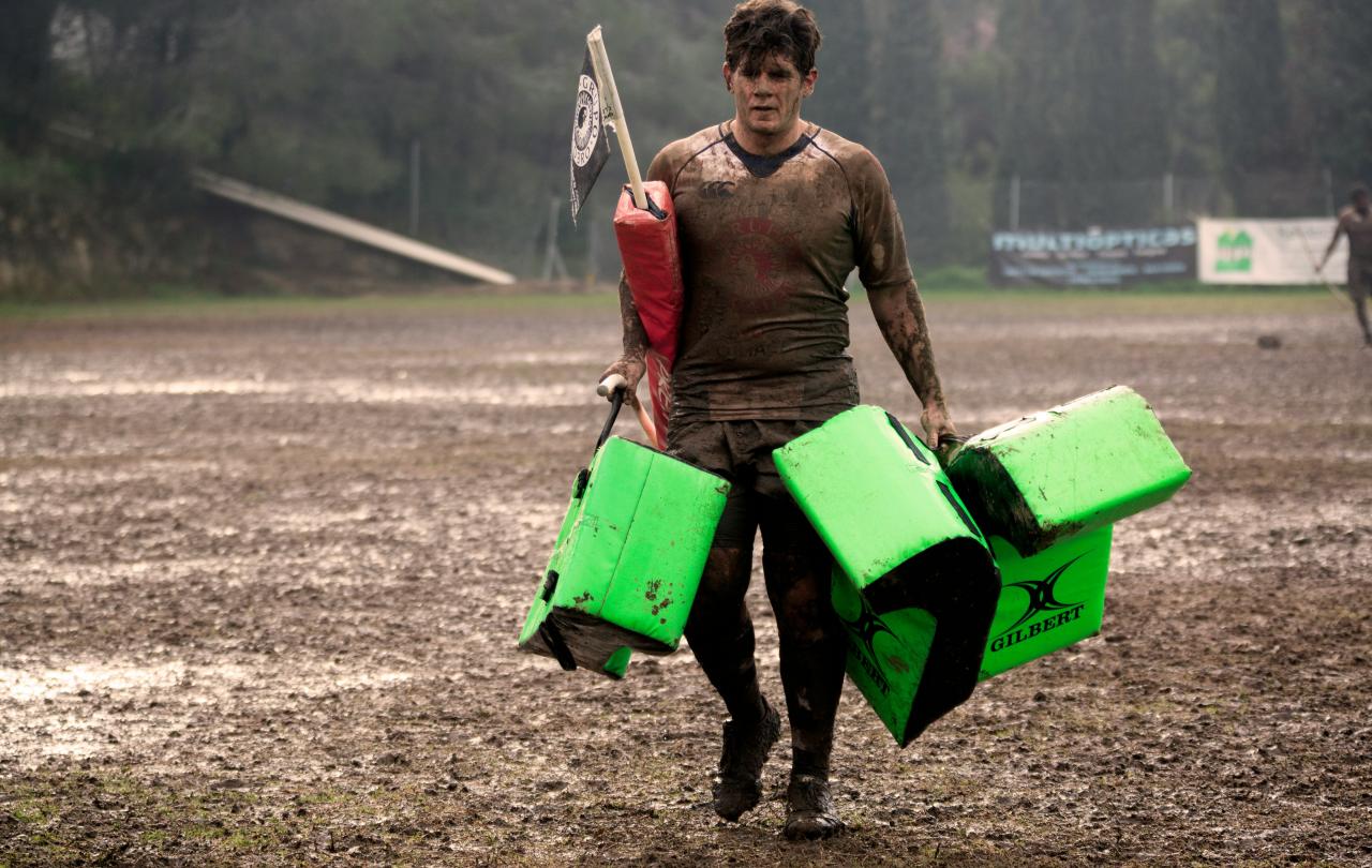 A muddy rugby player carries equipment off a pitch.