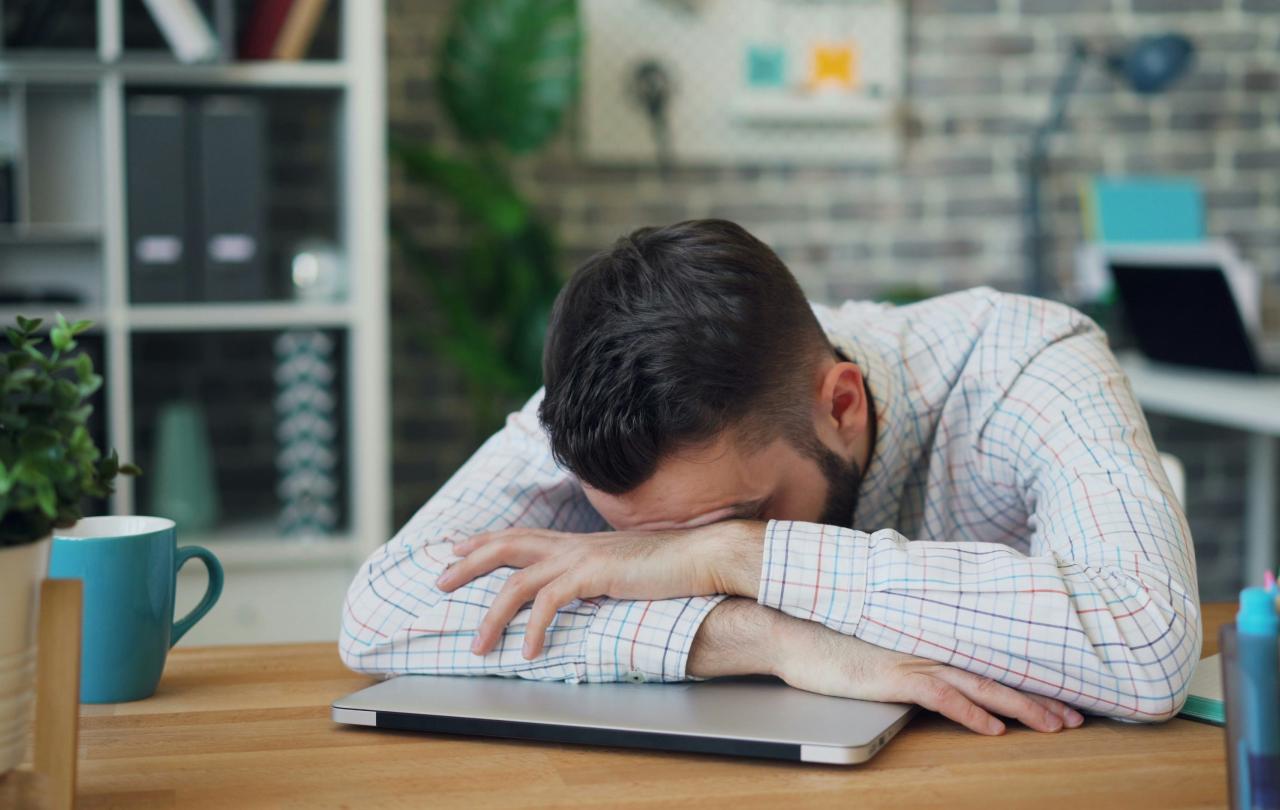 A man lies asleep on a closed lap top on a desk.