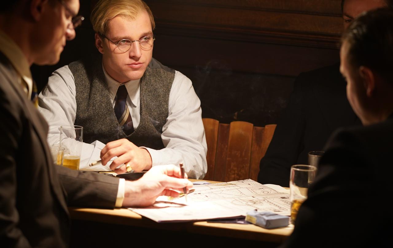 A man dressed in 1930s clothing, sits with others at a table looking pensive.