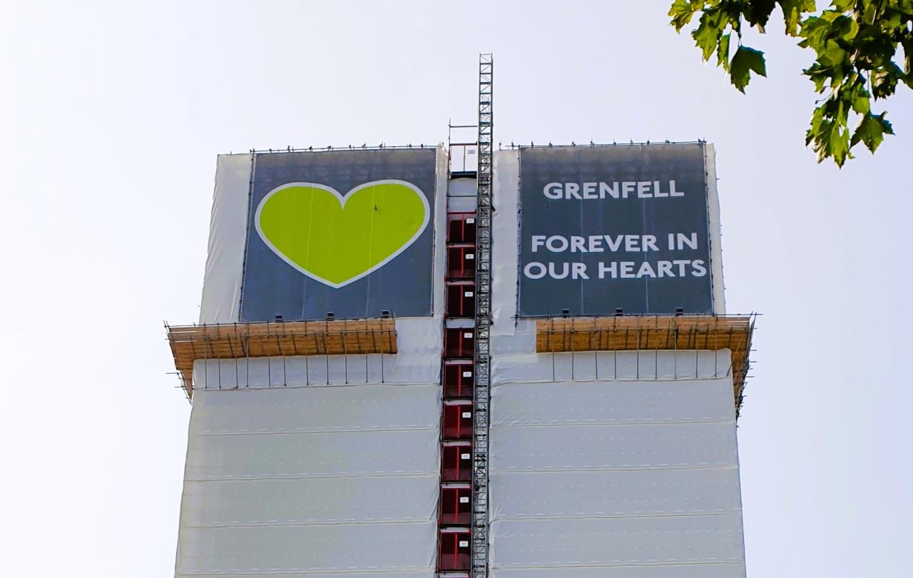 A white building wrap around a tower is topped by a green heart and the slogan: Grenfell Always In Our Heaets.
