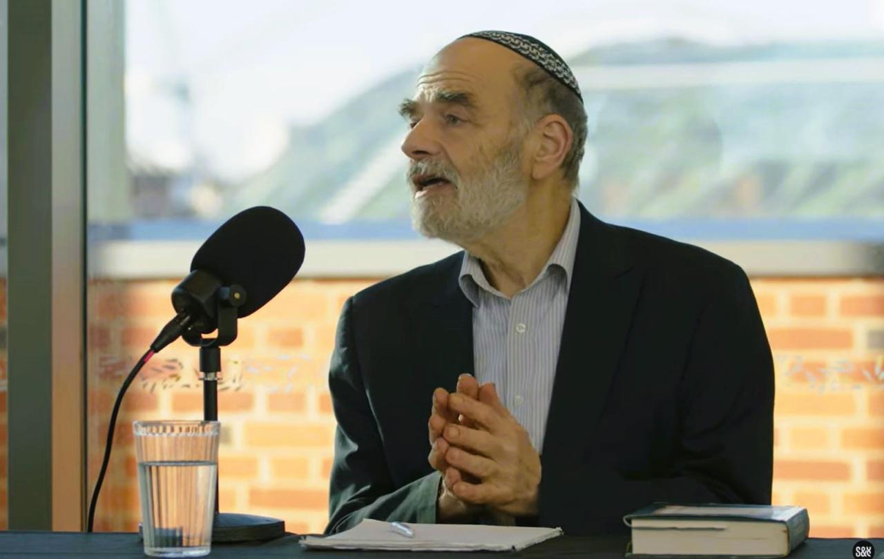 A podcast guest holds his hands together in front of himself as he raise his head in speech.