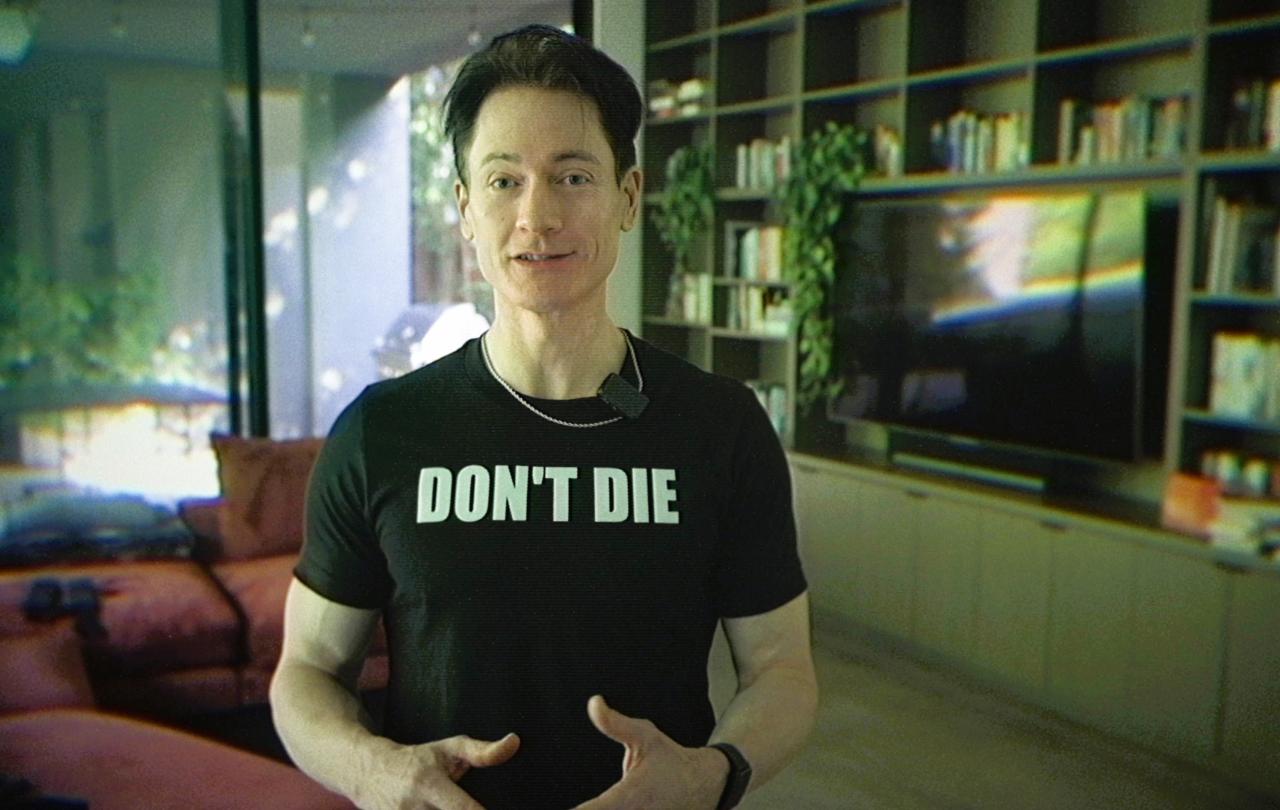 A man stands in his home wearing a black t-shirt that reads 'DON'T DIE'.