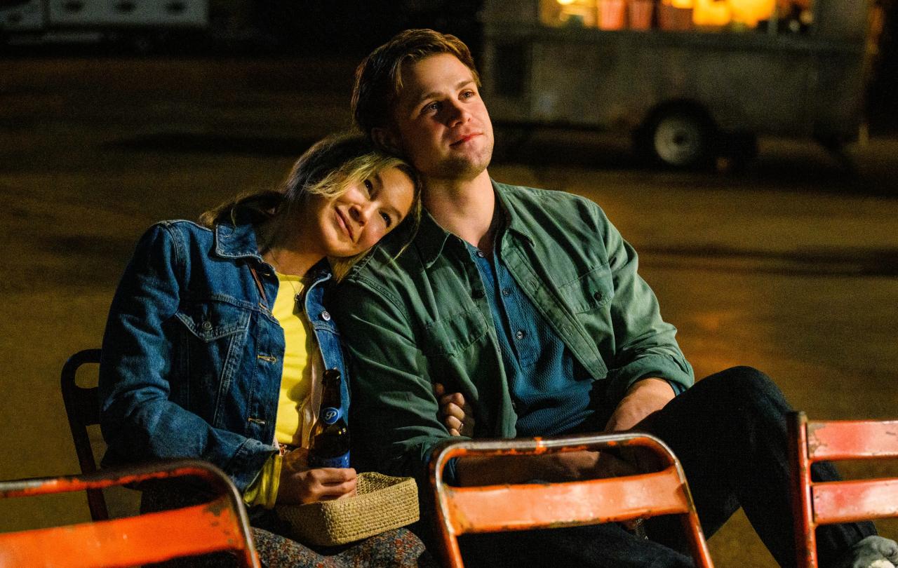 A couple sit on outdoor seats, her resting her head on his shoulder.