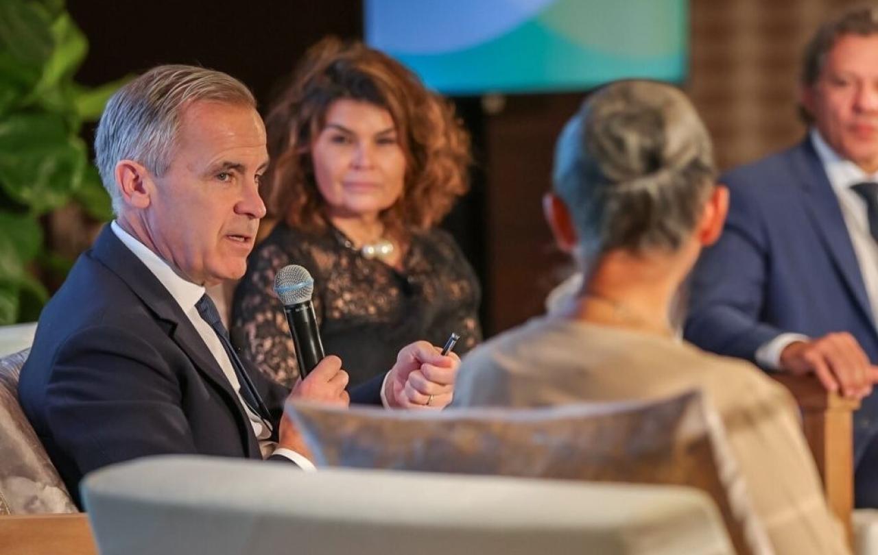 Mark Carney sits between two other speakers, holding a mic.