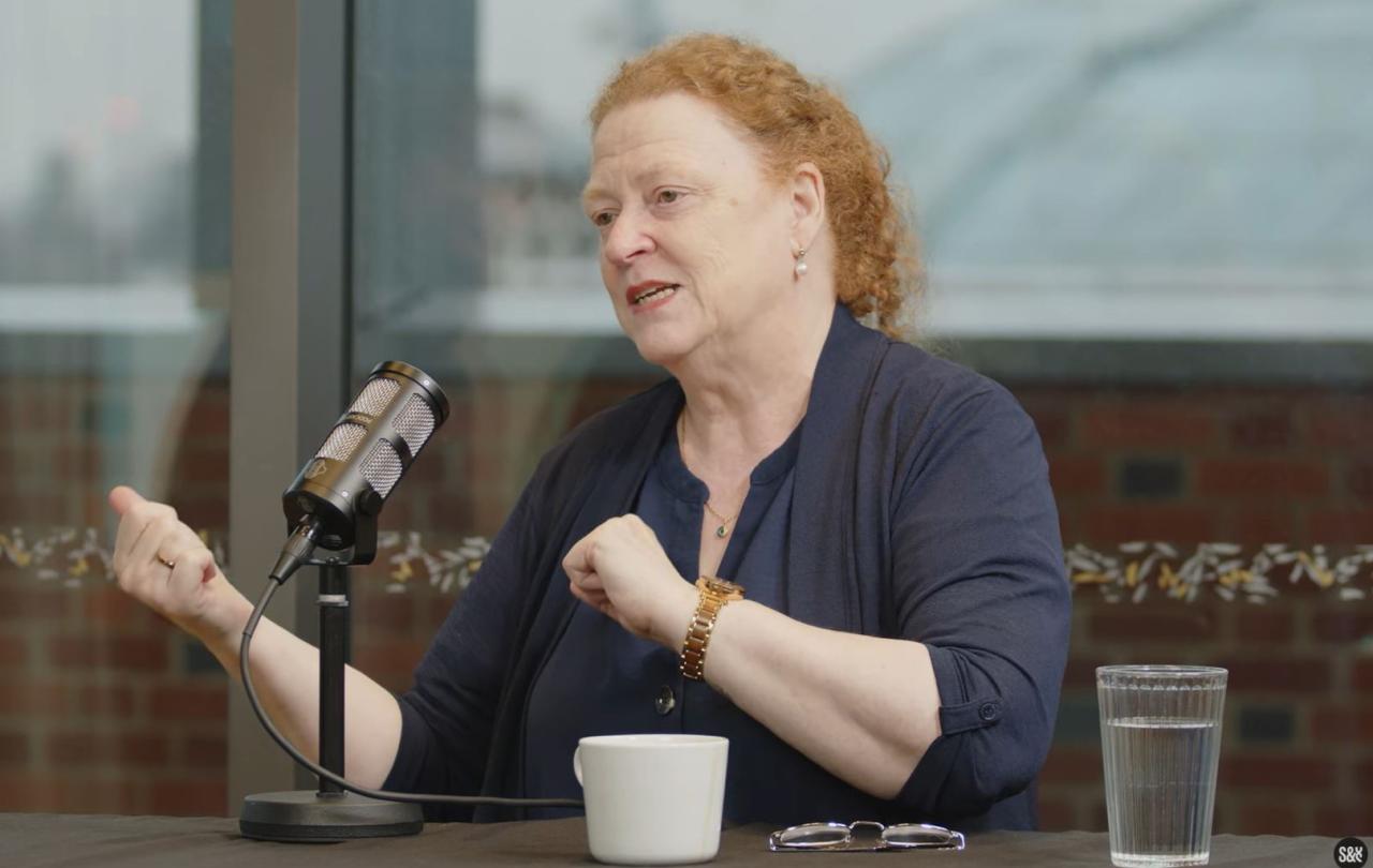 A woman guest on a podcast sits at a table with a mic in front, gesturing to the side with both hands.