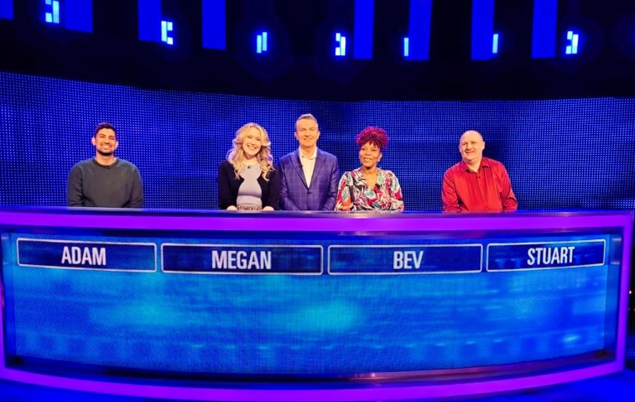 Four quiz show contestants stand behind a table with the show host.
