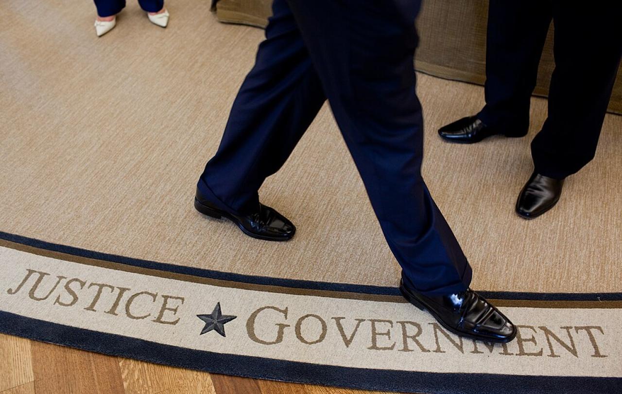 The feet and legs of someone walking on a white rug, beside the words Justice and Government woven in to it,
