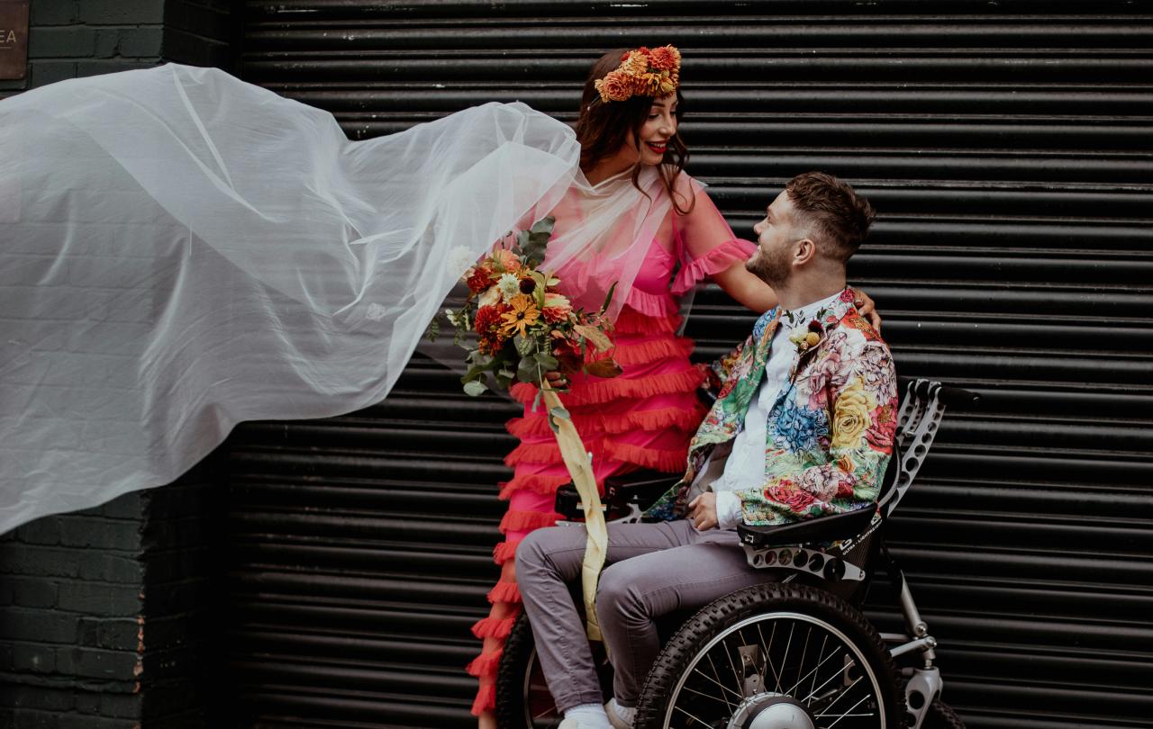 A bride dressed colourfully stands next to her groom, dressed similarly, as he sits in a wheelchair.