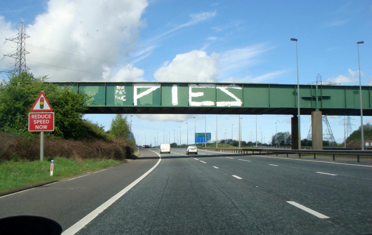 A green bridge spans a motorway, on its side is graffiti that reads 'PIES'