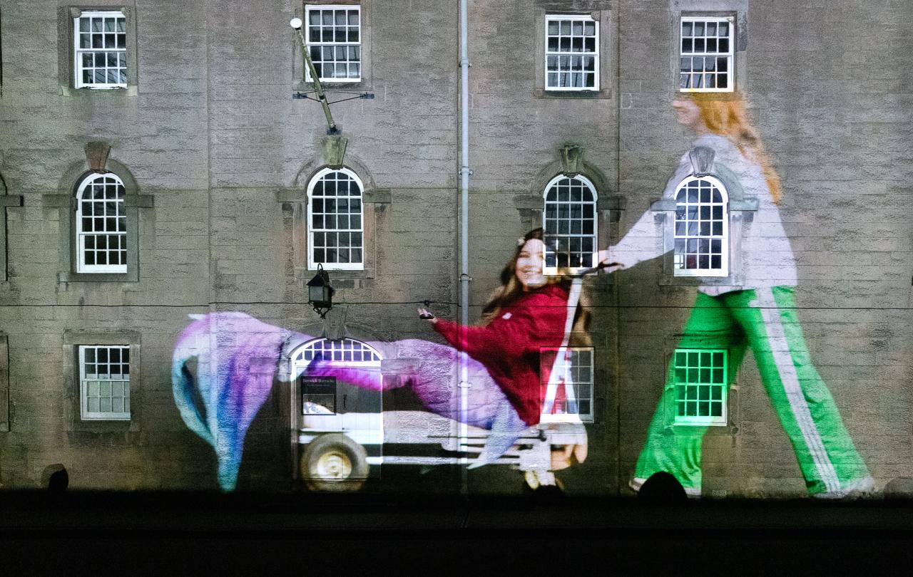 A projected image of a person dressed as a mermaid being pushed on a trolley falls on the wall and windows of an old barracks.
