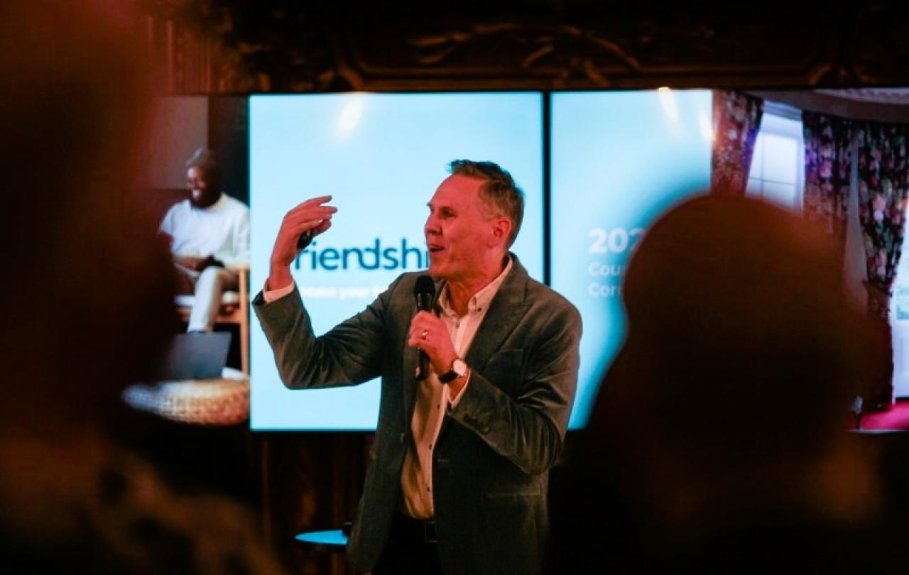 A speaker, standing in front of a screen, beckons with one hand, holding a mic with the other.