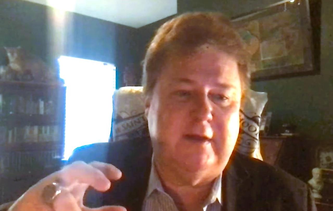 A podcast guest, on a screen, gestures with a raised hand.