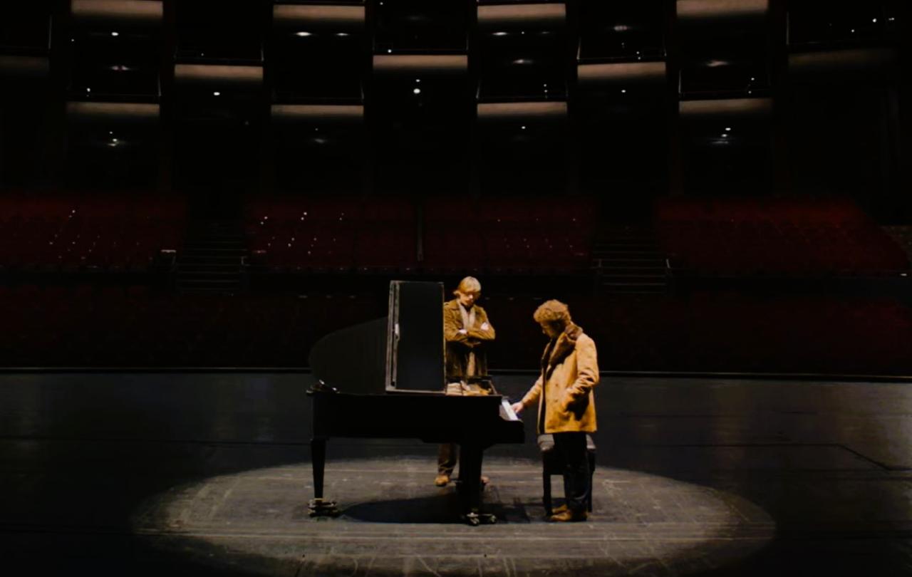 Two people sit and stand next to a grand piano on a stage.