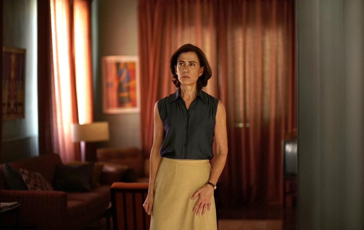A woman, wearing 70s clothing, stands, looking apprehensive.