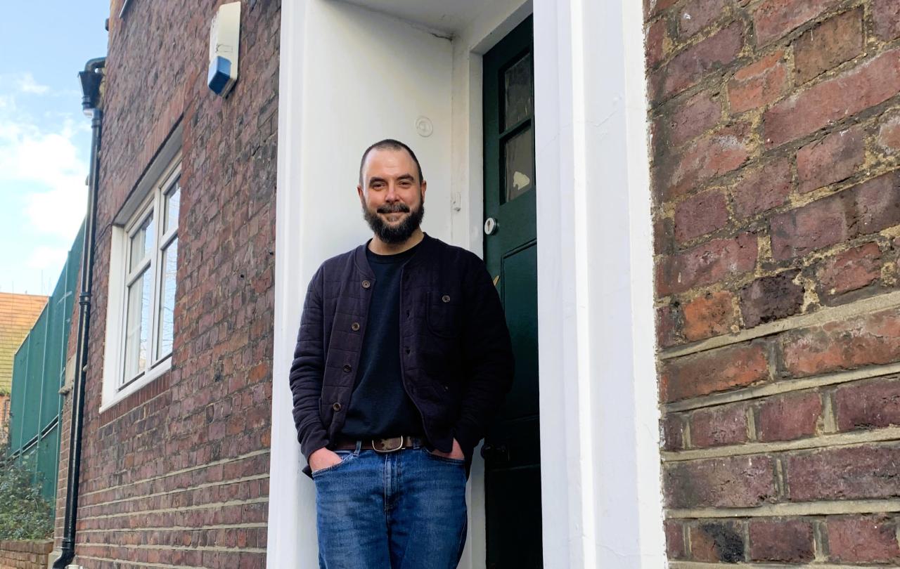 A man stands in a relaxed way on the doorstep of a brick building.