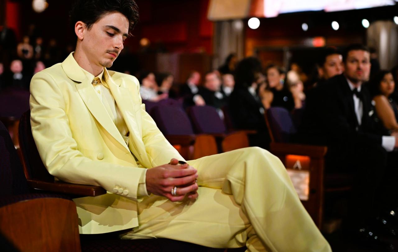 Timothee Chalamet, wearing light yellow suit reposes in a seat a the Oscars, clasping his hands, his eyes closed.
