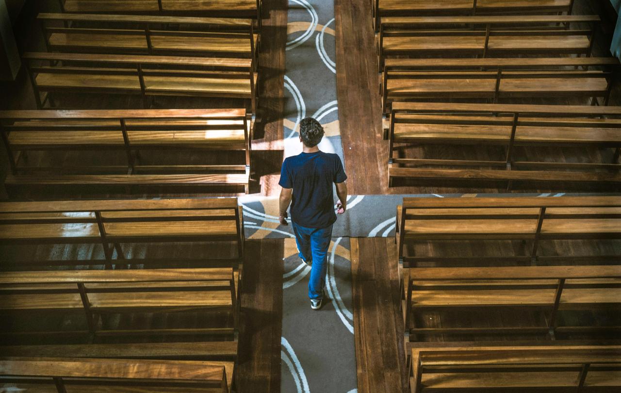 A casually dressed man strides down the aisle of a church between the pews.