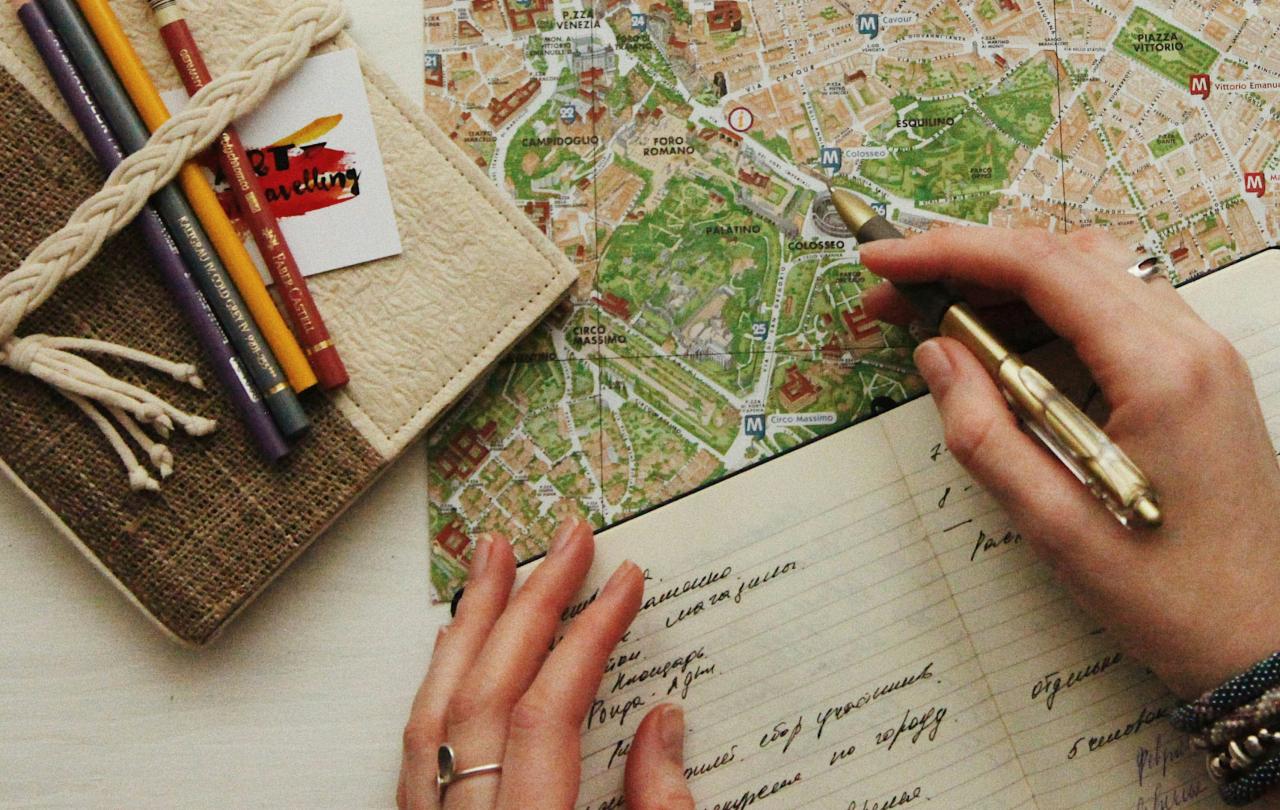 A hand holds a pen over a map, at the side is closed journal and colour pencils.