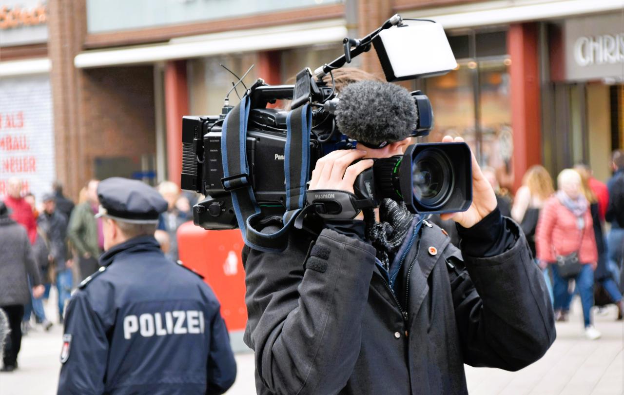 A news cameraman holding a camera, stands back to back to a police officer.