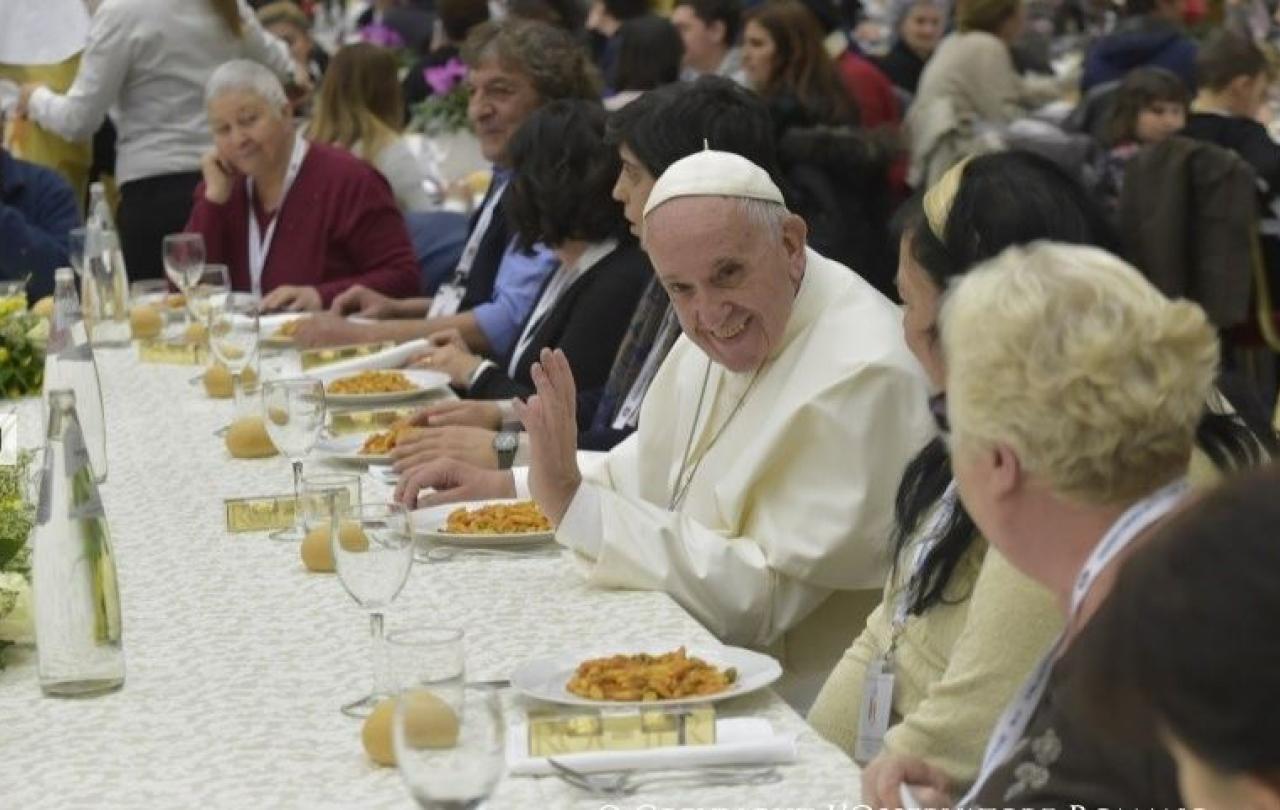 The Pope sits at a table having a meal, raising a hand of blessing
