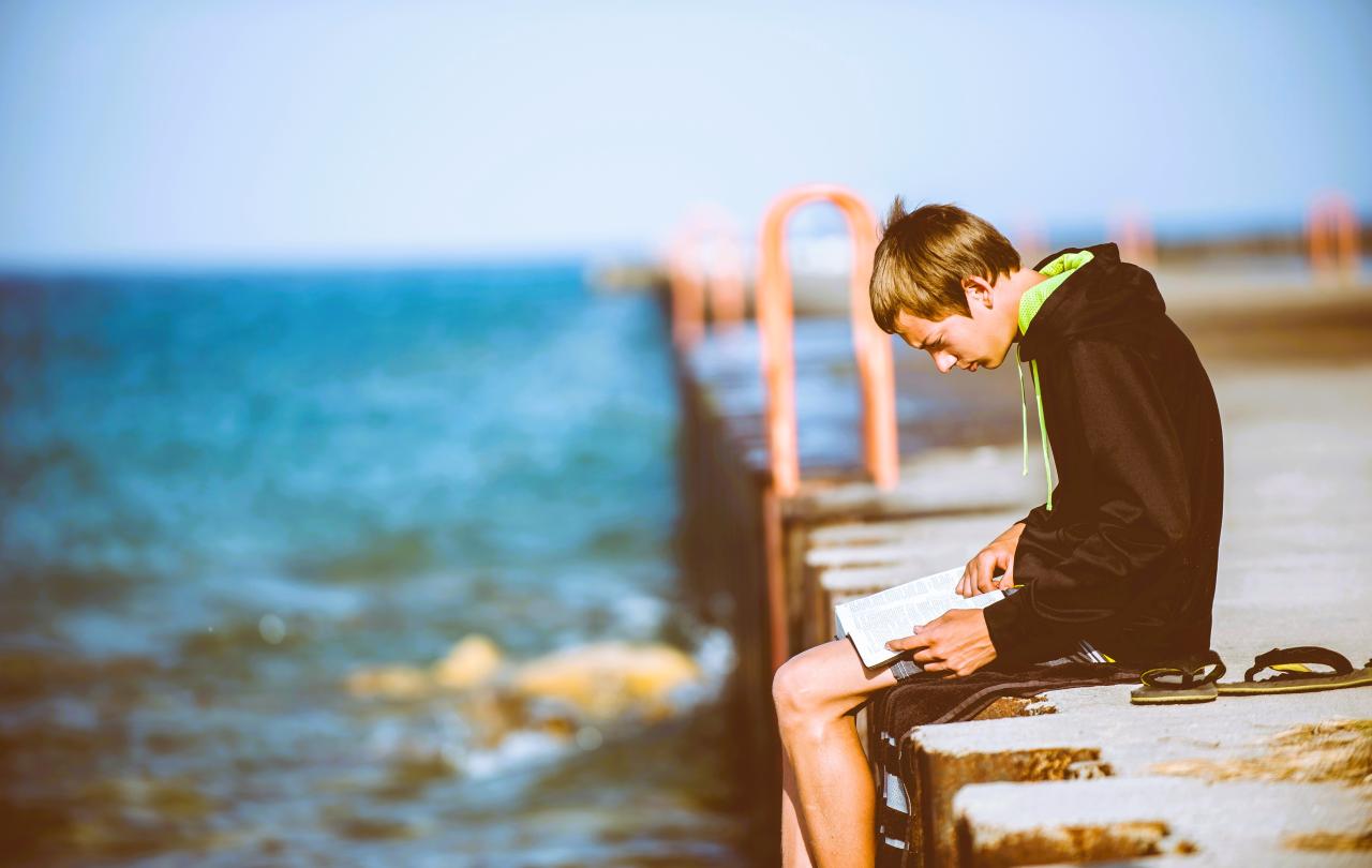 A man sits on a pier intently reading a book on his lap.