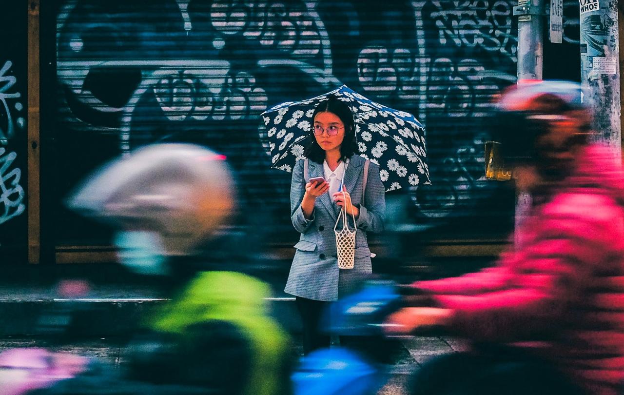 A woman stands across a busy roads, looking up from her phone in a sad way.