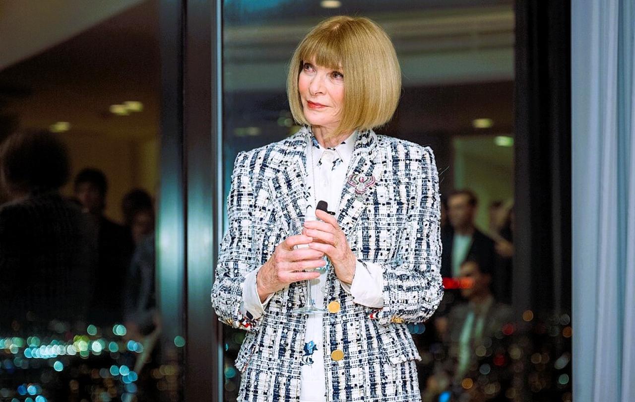 Anna Wintour stands holding a small mic.