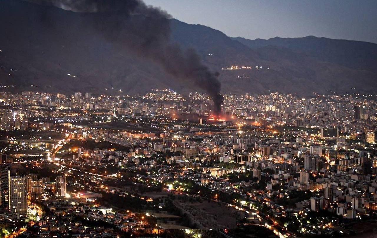 Smoke rises from the site of a bomb amid a high wide view of a city at twlight