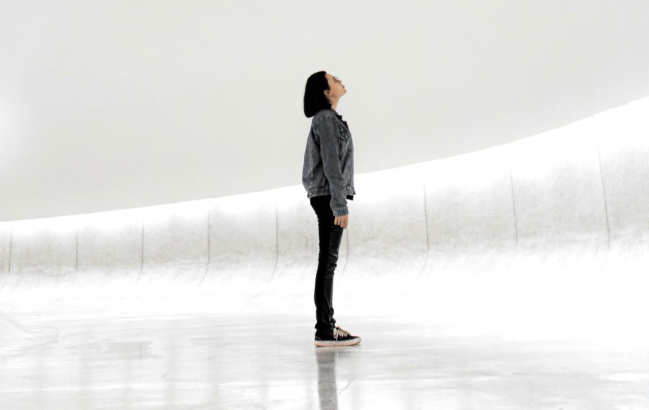 A woman stands against a sparse white background looking up.