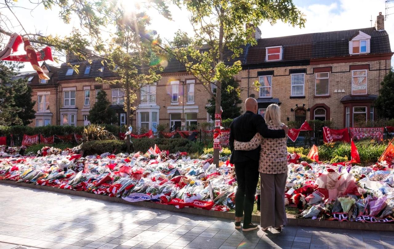 A couple hold each other as they look at floral tributes on the ground