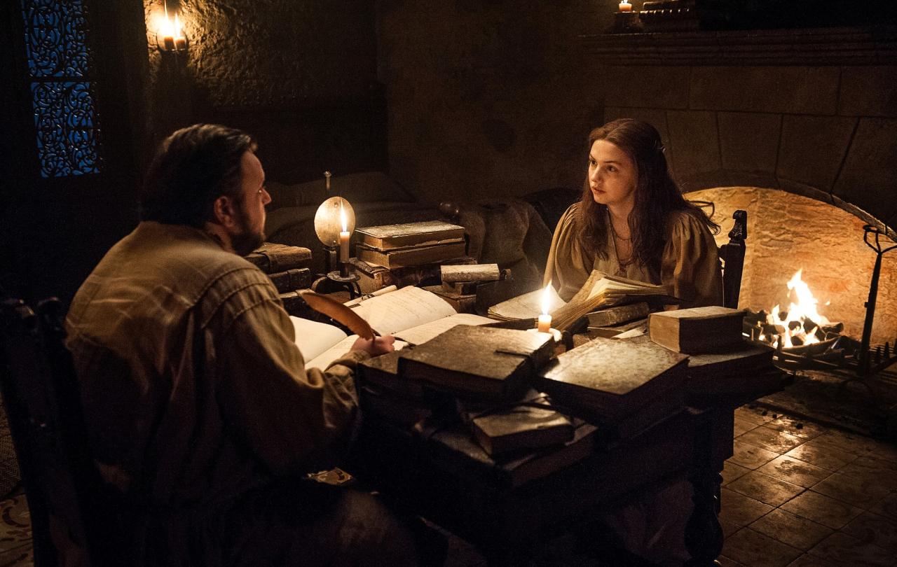 Two people sit at a table strewen with old books lit by candle light.