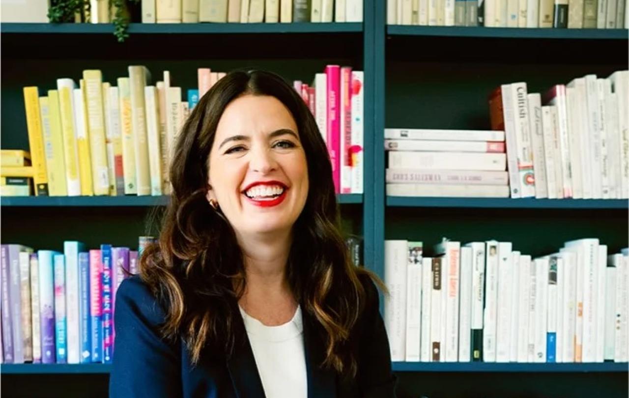 A woman sits and smiles in front of her bookcase.