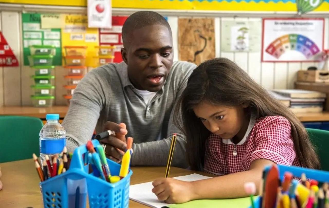 A teacher looks on as a young child concentrates on writing.