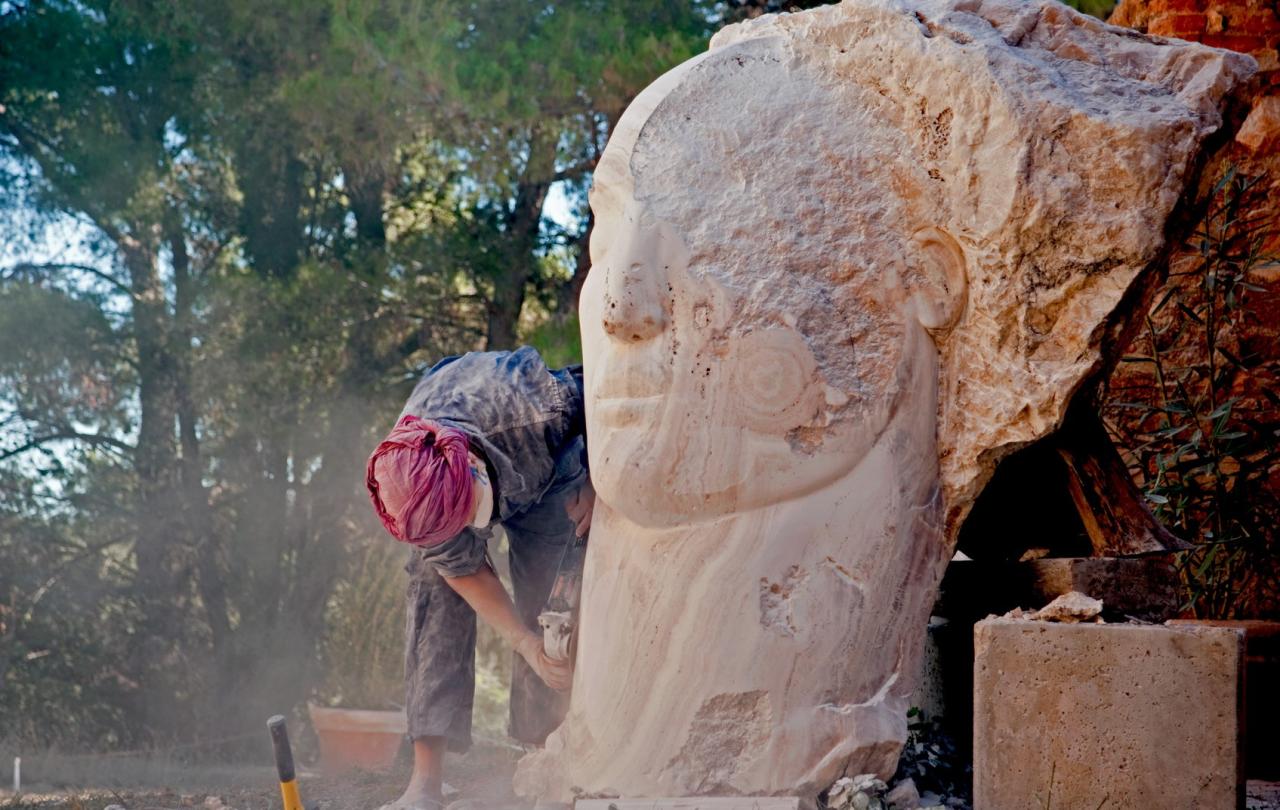 A sculptor works on a carved face emerging from a stone larger than her