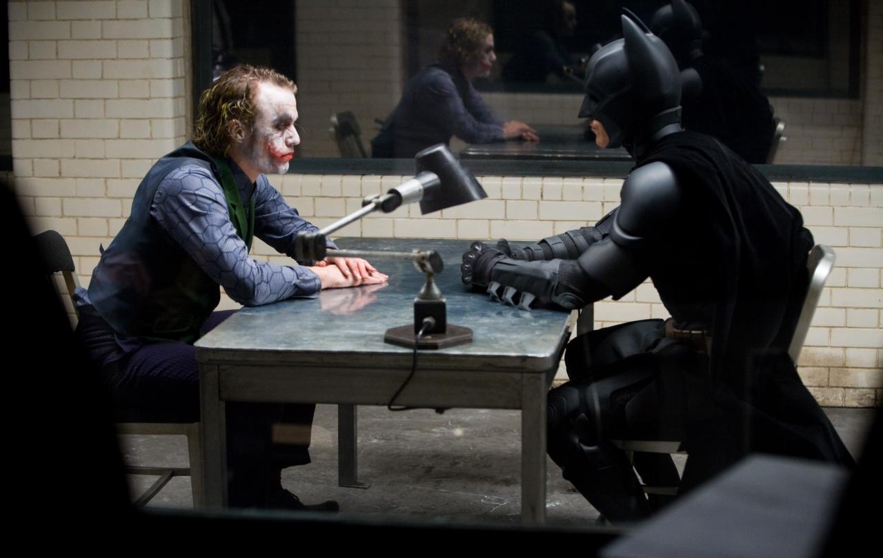 The Joker and Batman sit opposite each other in an interrogation room.