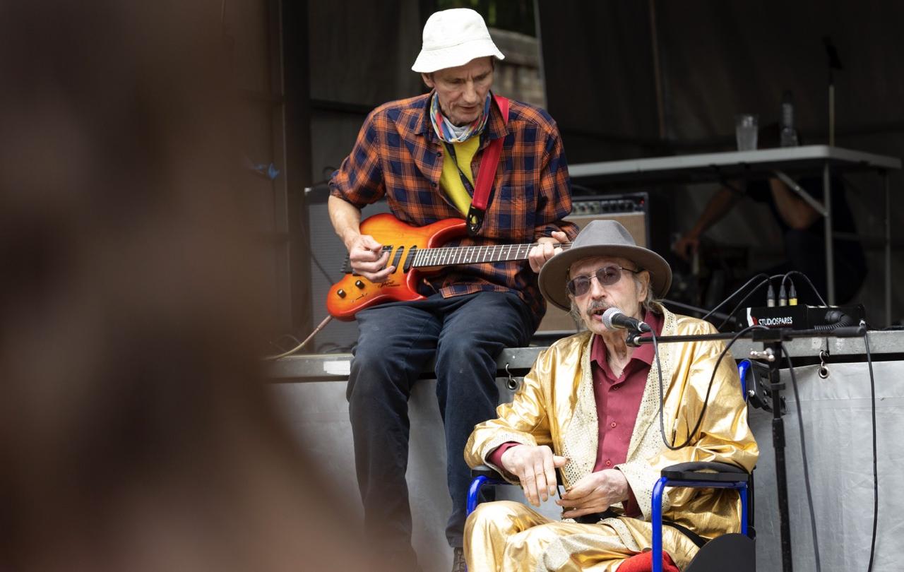 An elderly Elvis impersonator sits on a stage wearing a gold suit.