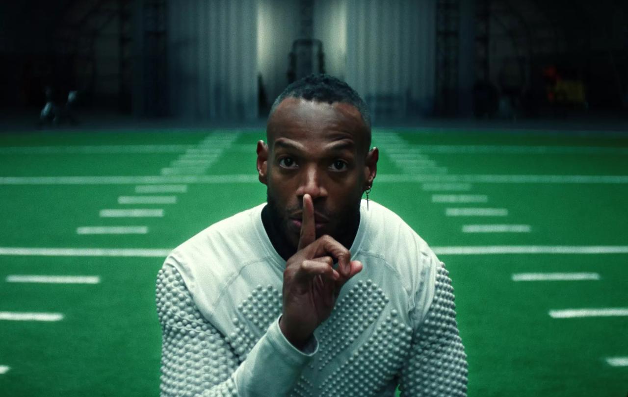 A player holding a finger to his lips stands in front of an indoor American football pitch.