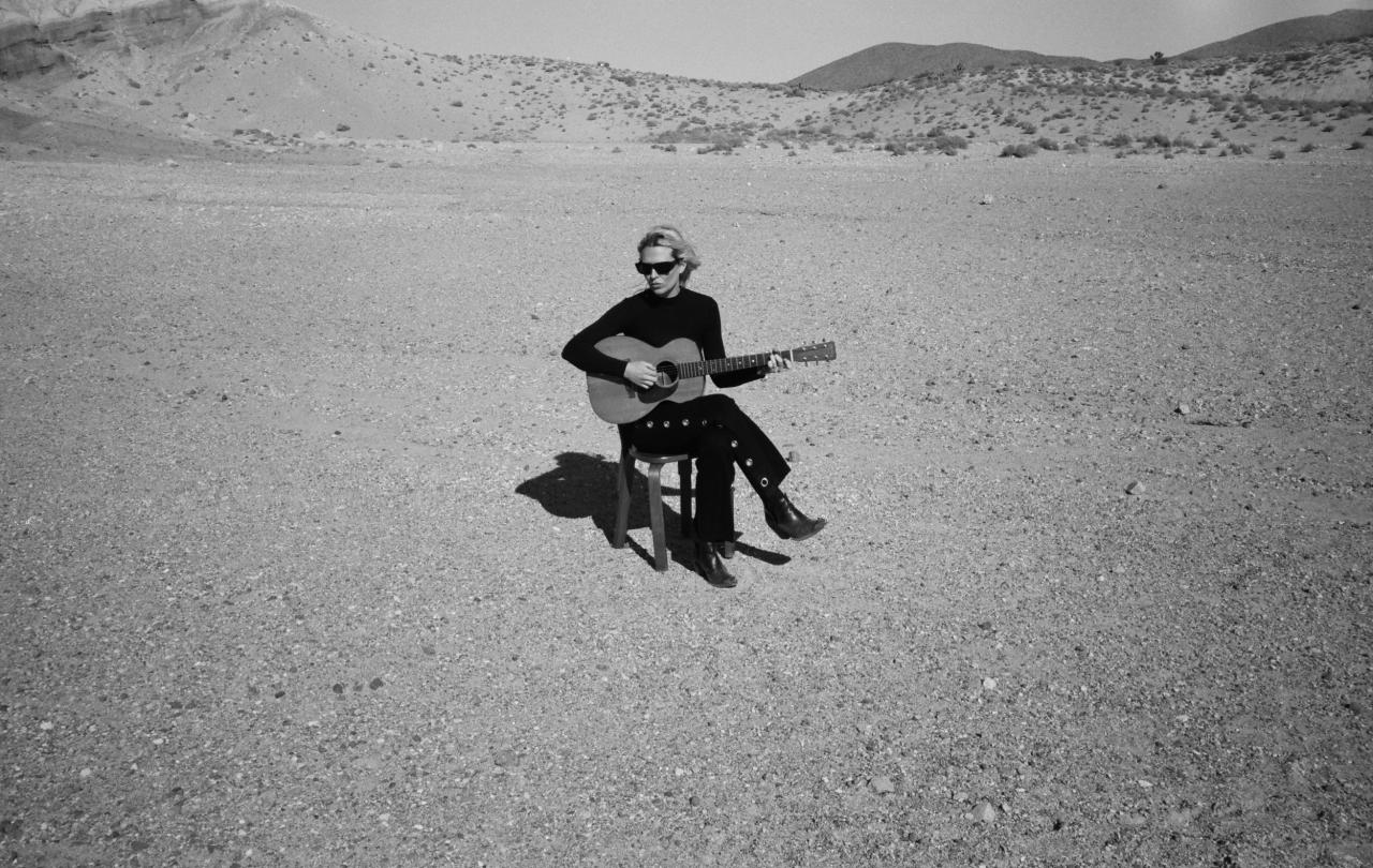A musician wearing black sits on a chair in a desert holding her guitar.