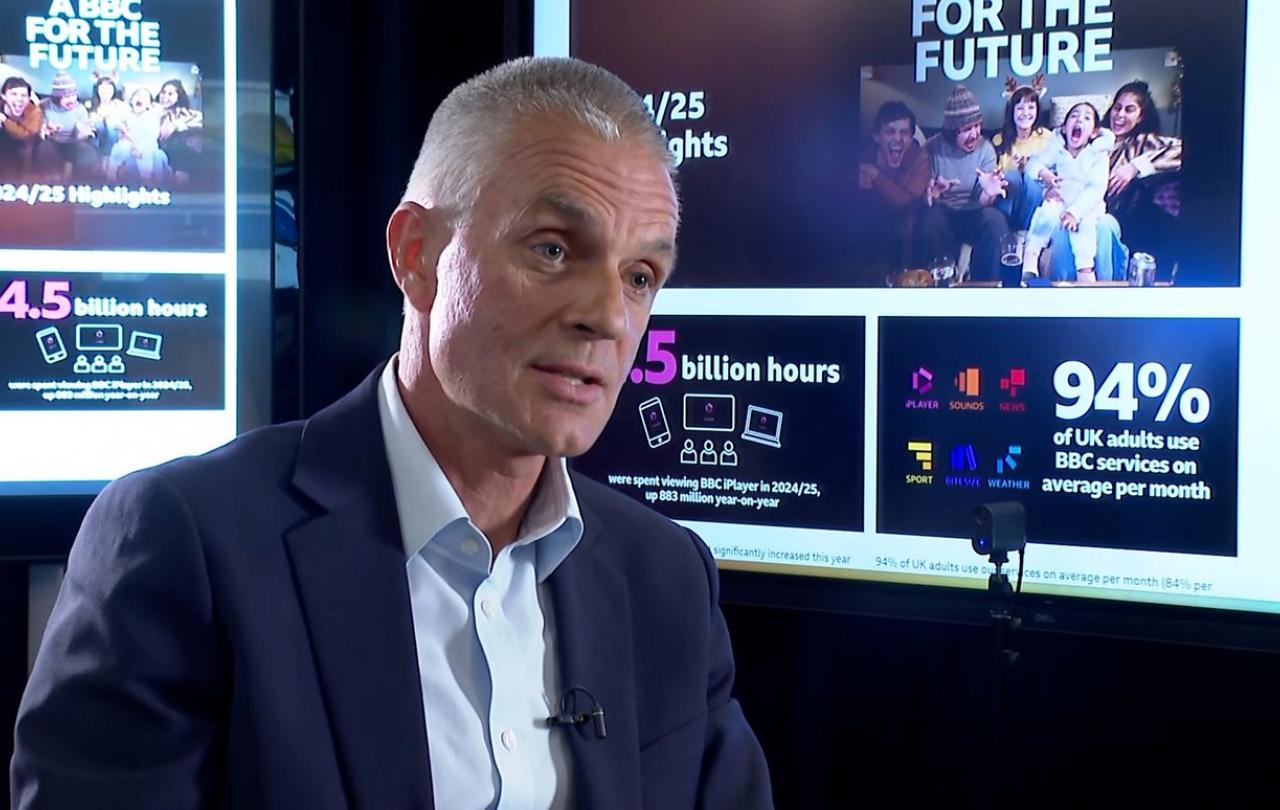 TIm Davie, sits in an interview in front of screens showing facts about the BBC