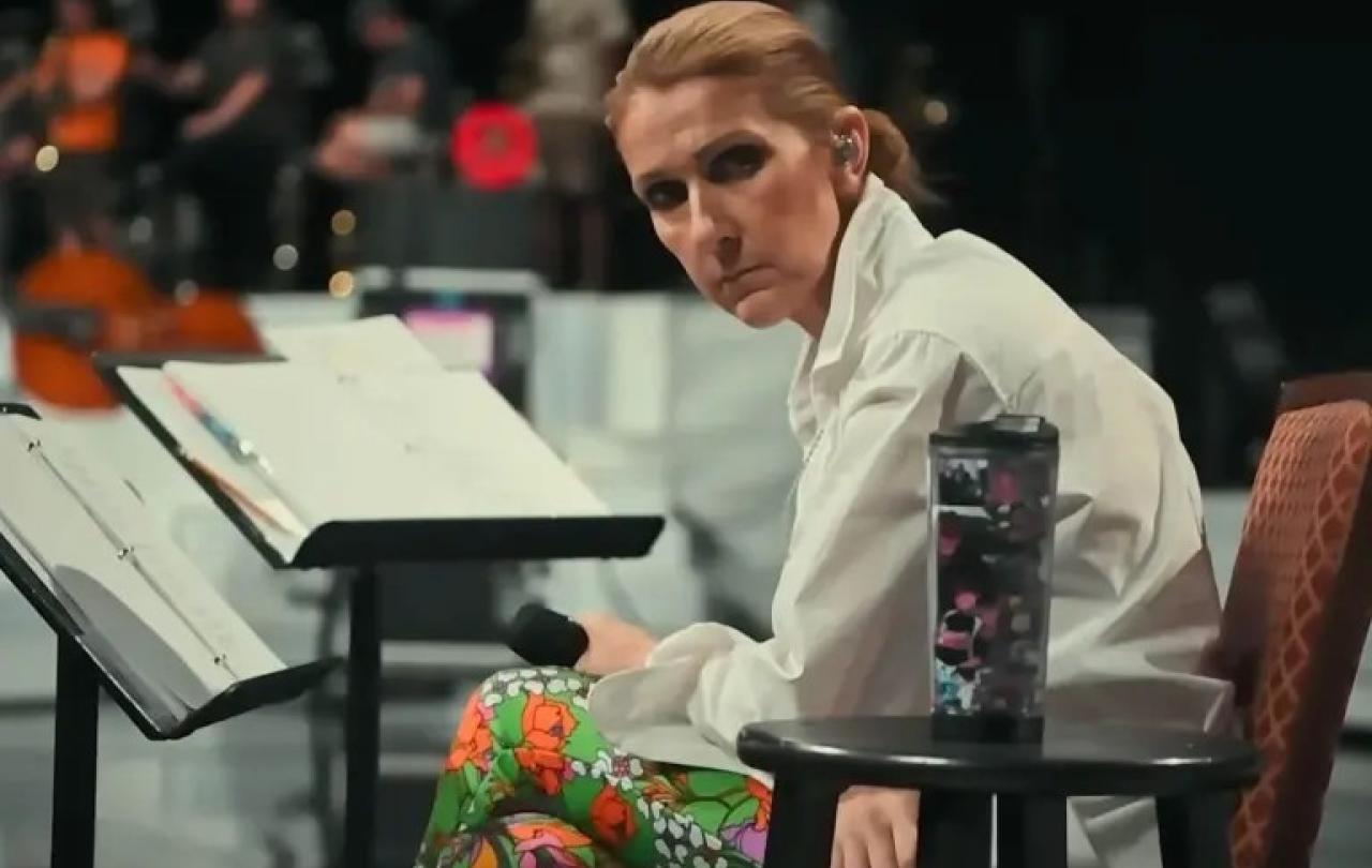 A seated Celine Dion, leans forward, head to the side, holding a mic.